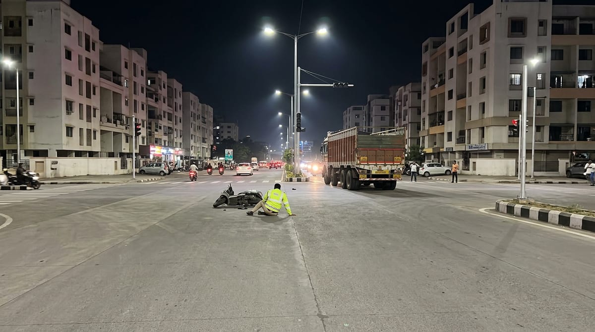 At night, a scooter lies on the road with a person sitting next to it, a truck and buildings in the background.